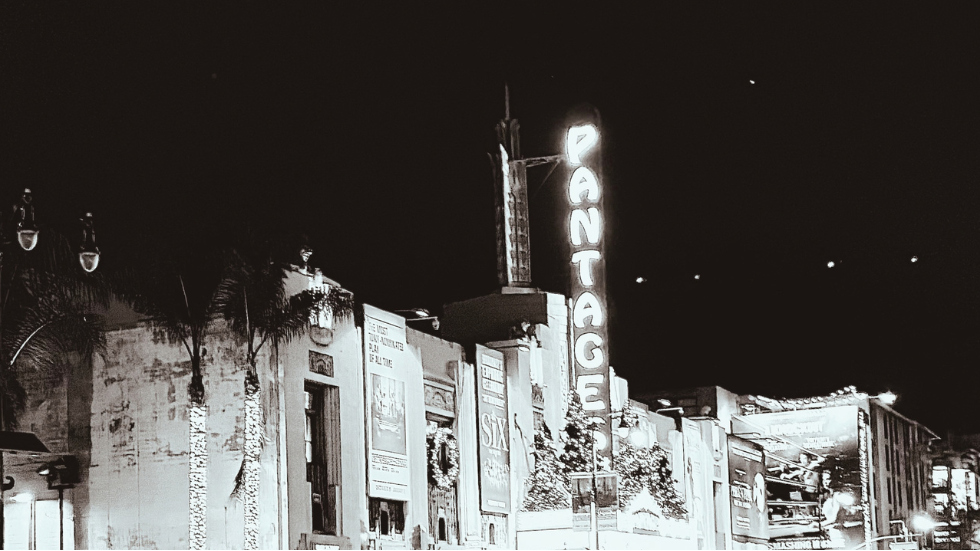 Black and white night photograph of a city street with parked cars, storefronts, and a tall illuminated theater sign reading 'Grand' or similar.